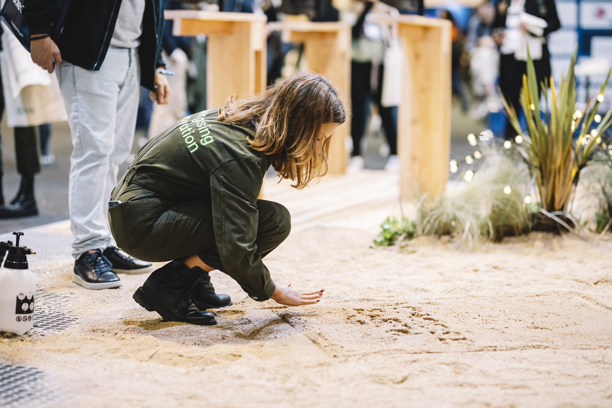 A person squats on a sandy surface. Their right hand is splayed inches above the ground, where something appears to be drawn or written in the sand. People appear to be walking by in the background. This is the exhibition floor at Web Summit.