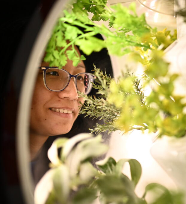 A smiling person seen through a round window. Their face is partially blocked by a variety of leafy plants.