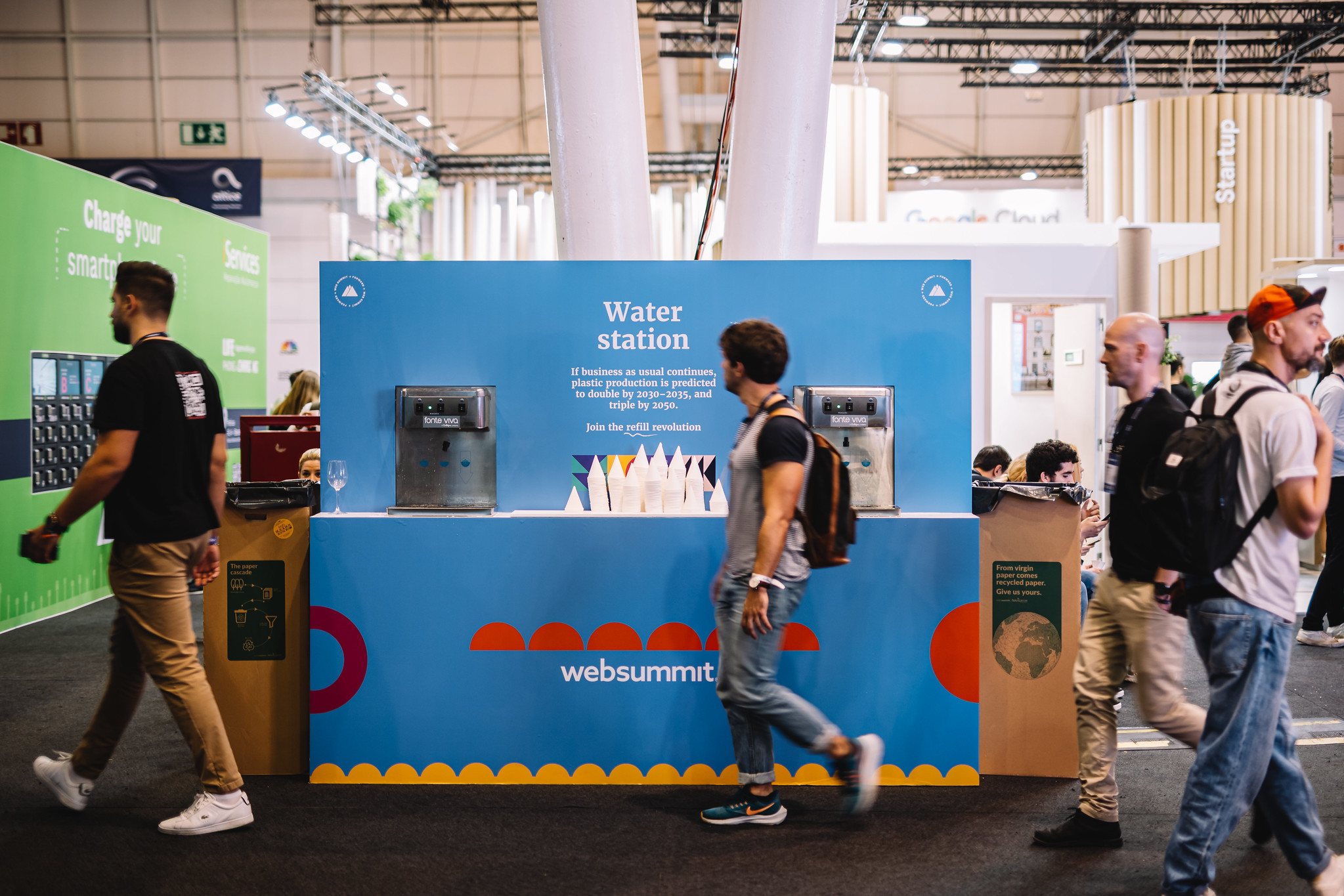 A person walks past a water station with Web Summit branding on it. The water station is on an exhibition floor with high ceilings and industrial lighting.